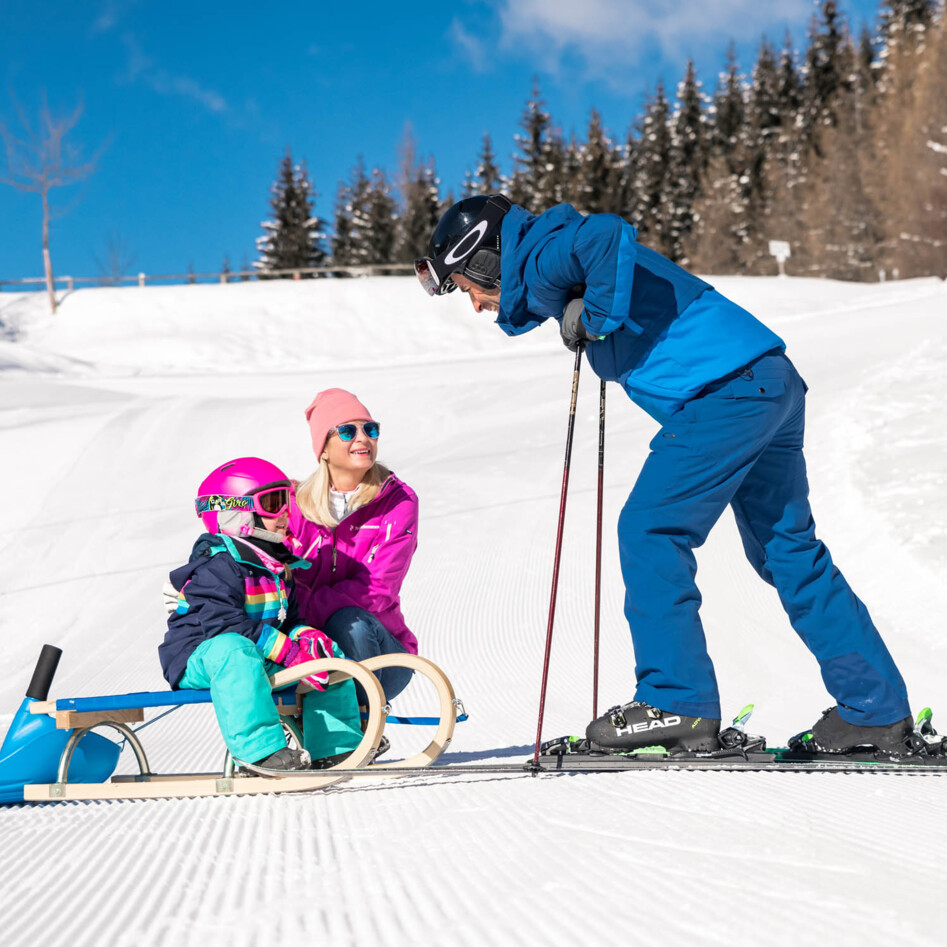 Eine Familie beim Rodeln und Skifahren im Schnee an einem sonnigen Wintertag.
