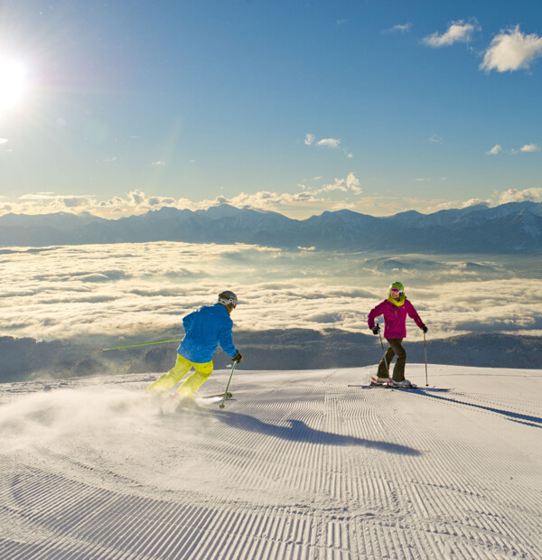 Two skiers carve down a freshly groomed slope above a sea of clouds, with sunlit Alpine peaks stretching across the horizon.