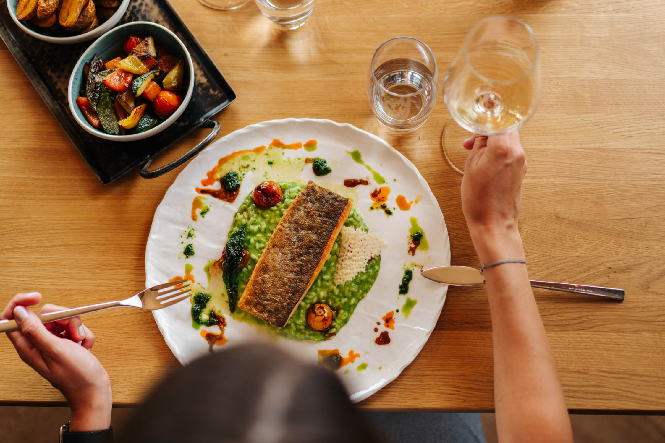 Eine Person genießt ein Gericht mit Fisch und Gemüse, begleitet von einem Glas Weißwein.
