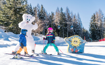 Zwei Kinder lernen Skifahren mit dem Maskottchen der Region Bino Bär. Im Hintergrund sind verschneite Bäume und Skilifte zu sehen.