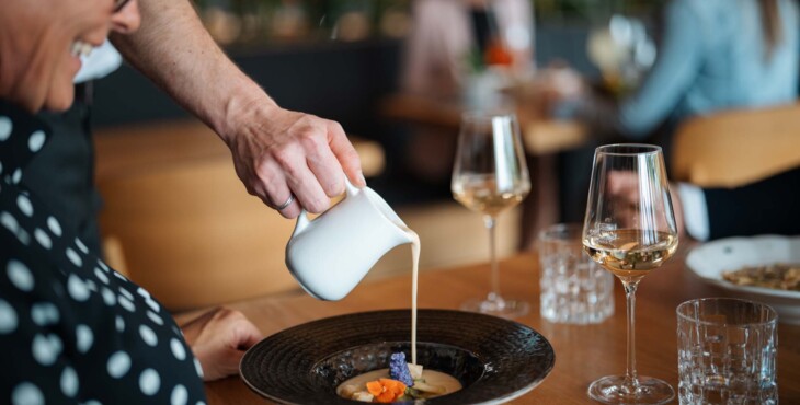Ein Kellner gießt Soße über ein Gericht, während Gäste sich auf ihr bestelltes Gericht freuen.