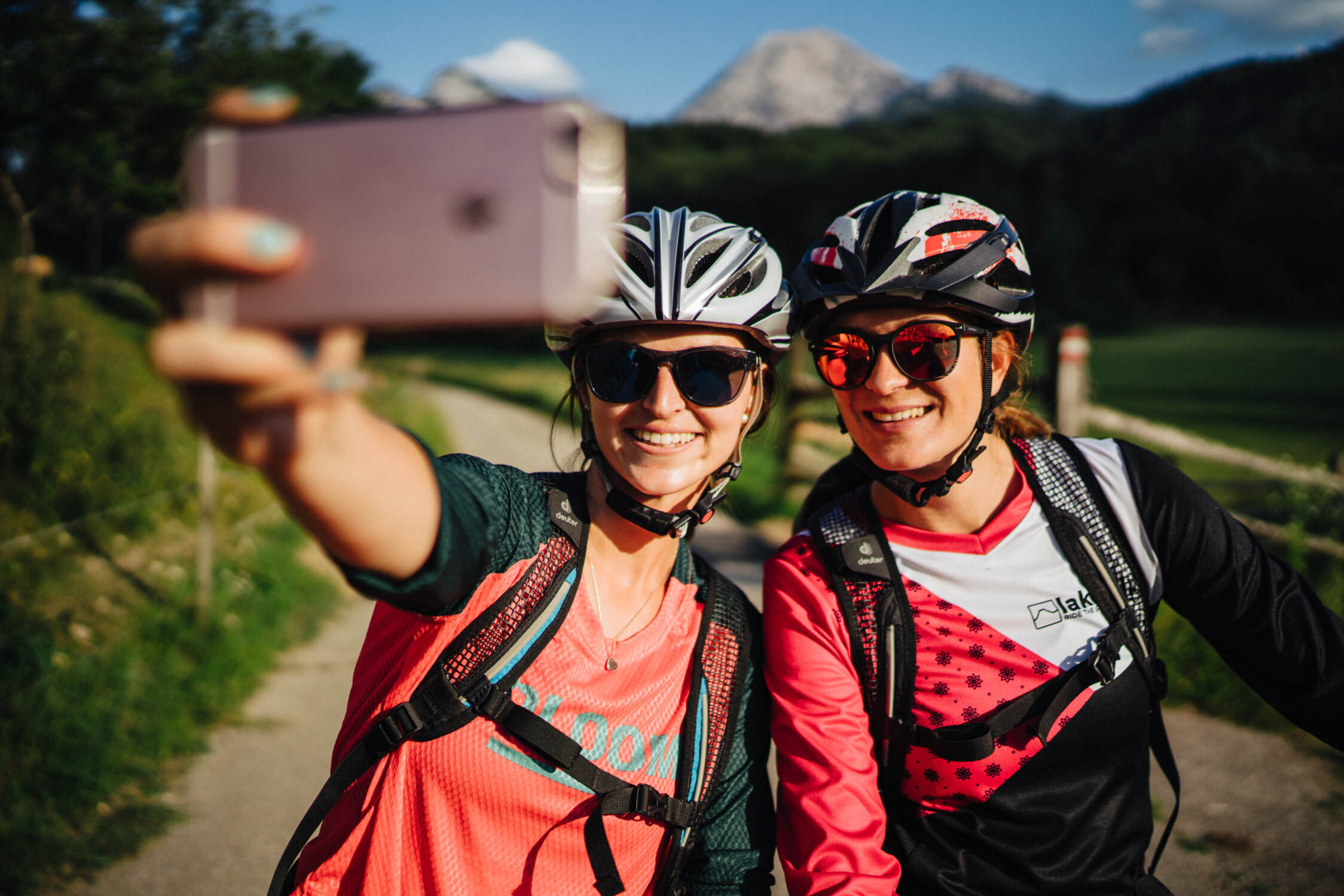 Zwei Radfahrerinnen machen ein Selfie auf einem sonnigen Weg mit Bergen im Hintergrund.