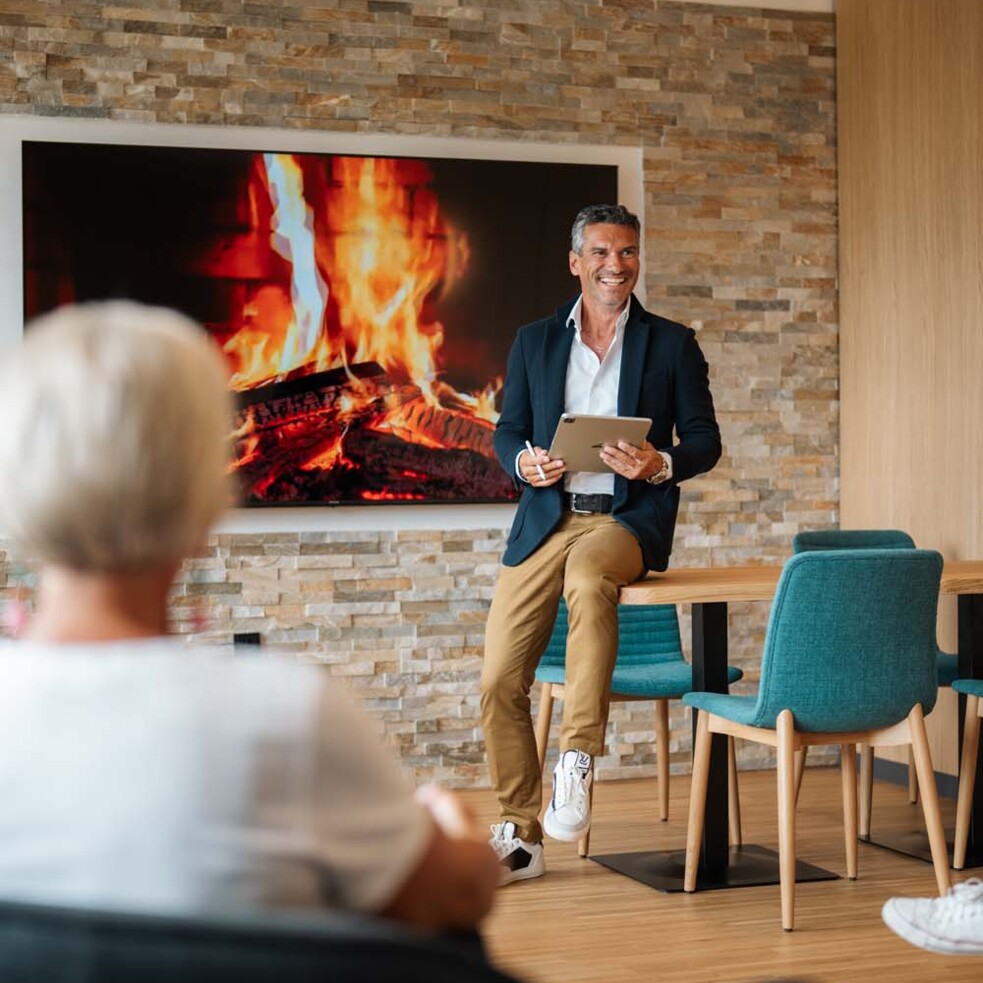 Ein Mann hält eine Präsentation in einem gemütlichen Raum mit moderner Einrichtung. Auf dem Bildschirm wird Kaminfeuer angezeigt. 