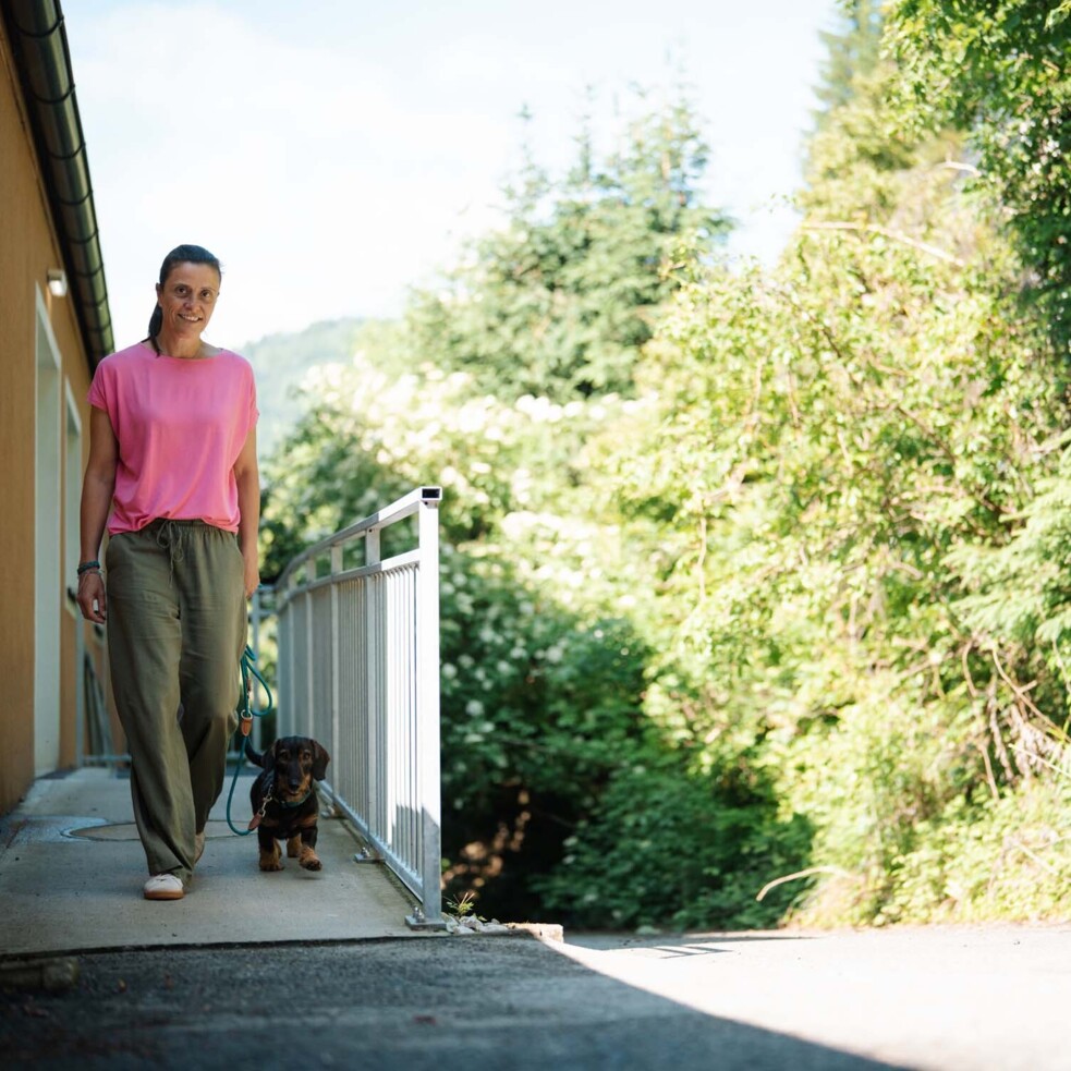 Eine Frau in rosa T-Shirt spaziert mit einem Hund an der Leine entlang eines Weges, umgeben von grüner Natur.