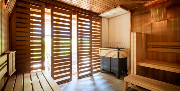 Innenansicht einer modernen Holzsauna mit Bänken und Ofen, durch Lamellen dringt Tageslicht ein.