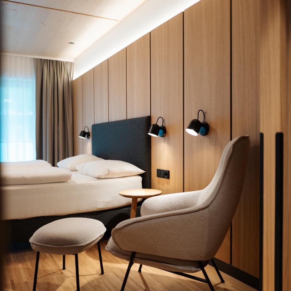 Modern hotel room with wooden paneling, a comfortable armchair, ottoman, and a bed. Warm lighting enhances the cozy atmosphere.