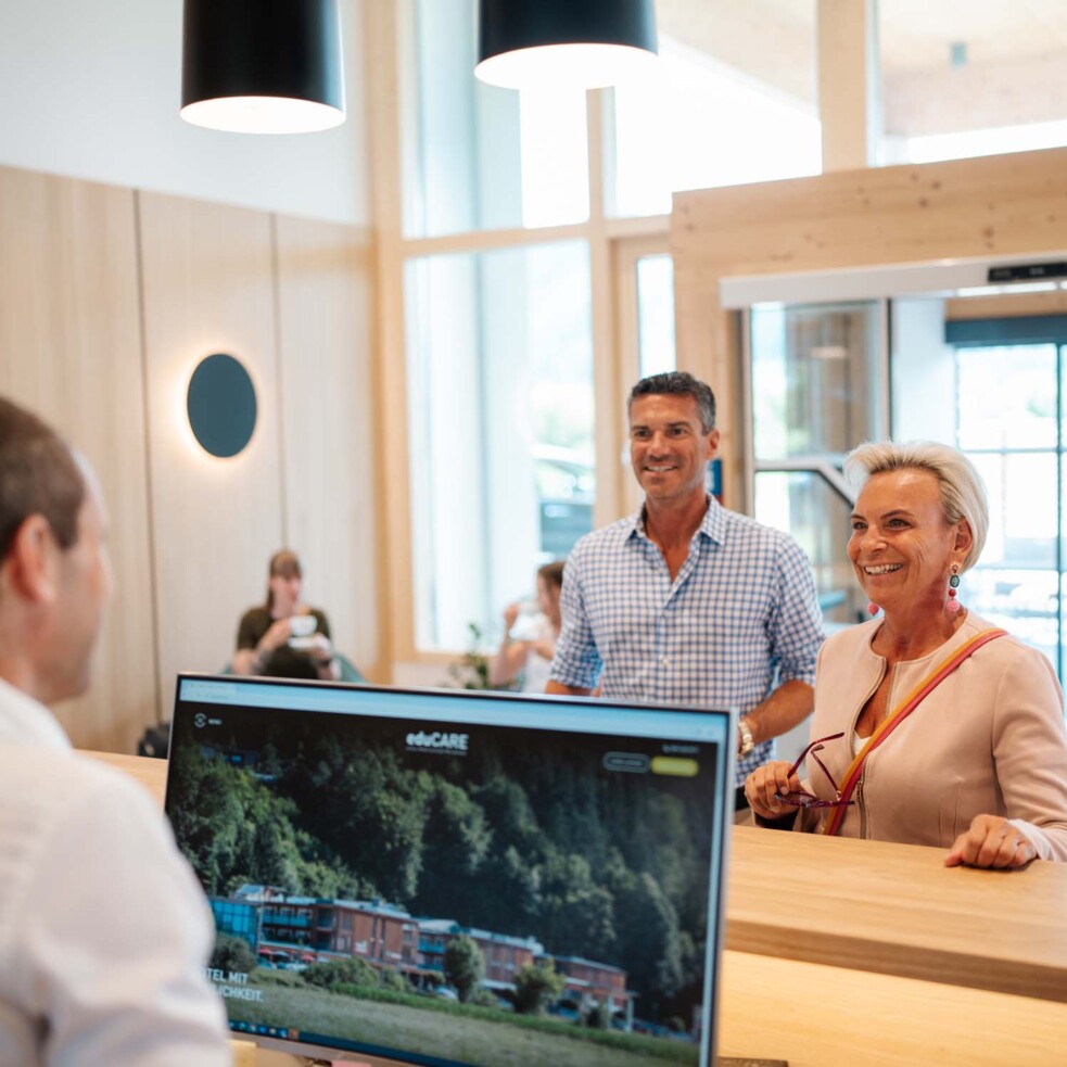 A couple checking in at the reception of the Hotel Educare. The couple smiles at a member of staff in a modern, bright hotel lobby.