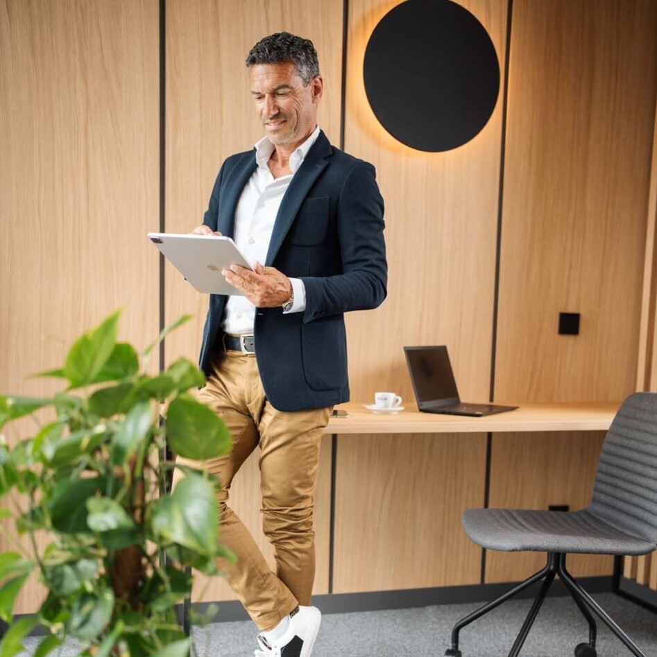 A man in a blazer stands in a modern office, holding a tablet. A plant and laptop are visible, with wooden paneling in the background.