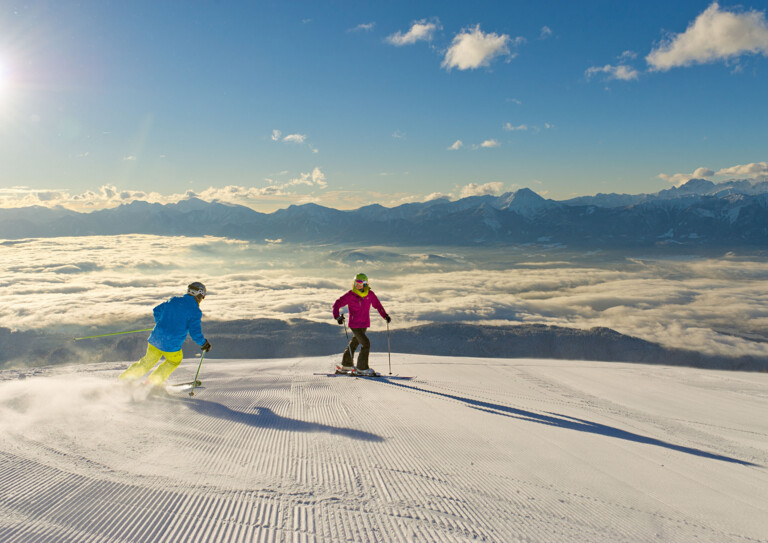 Zwei Personen beim Skifahren auf einer Piste mit Ausblicks auf eine Wolkendecke über dem Tal bei Sonnenschein