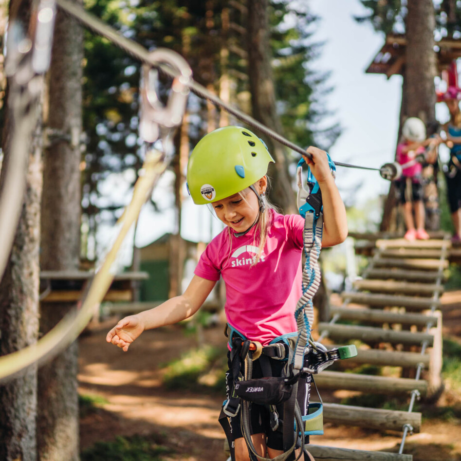 Ein Kind mit Schutzhelm balanciert auf einem Hochseilgarten im Wald. Im Hintergrund sind weitere Personen beim Klettern zu sehen.