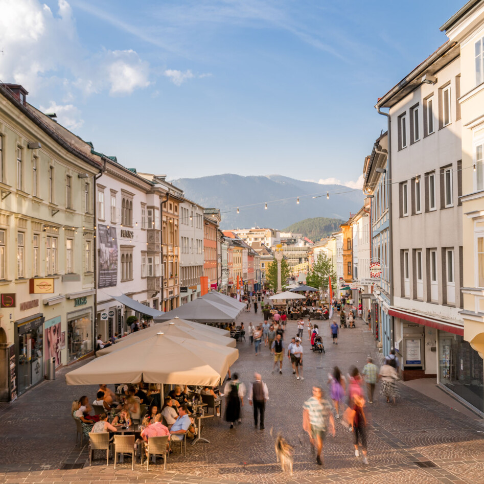 Belebte Einkaufsstraße in der Villacher Innenstadt mit Cafés und Geschäften, umgeben von historischen Gebäuden und Bergen im Hintergrund.