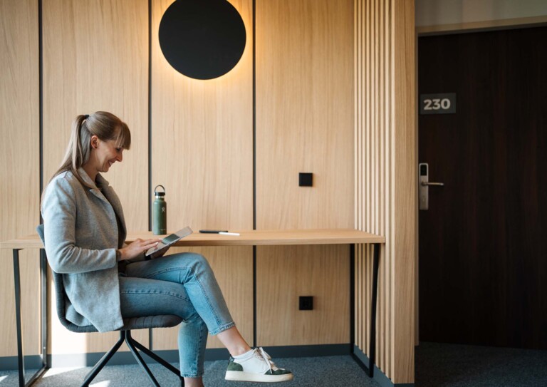 Eine Frau sitzt in einem modernen Hotelzimmer und liest auf einem Tablet, neben ihr steht eine Wasserflasche auf dem Tisch.