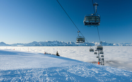 Sessellift über schneebedecktem Berg mit Blick auf Wolkenmeer und Alpenpanorama.