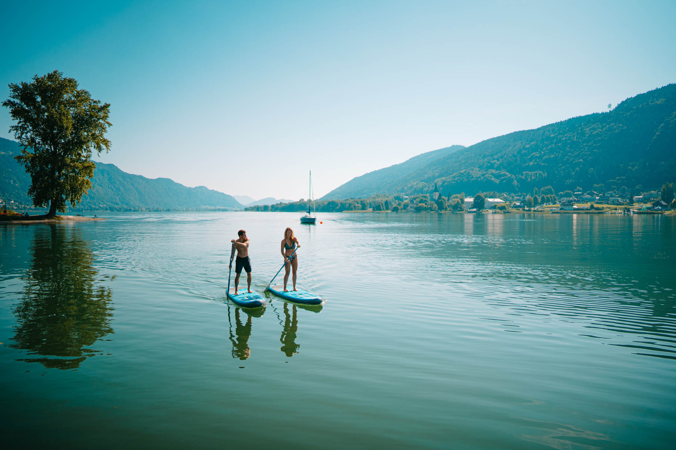 Two people paddleboarding on the Ossiacher See, mountains and trees in the background.