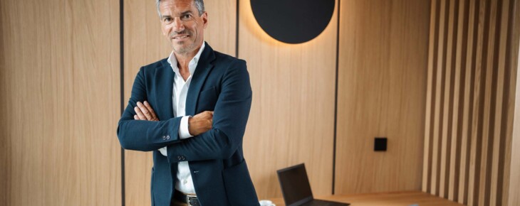 Ein Mann steht mit verschränkten Armen vor einem Schreibtisch mit Laptop.