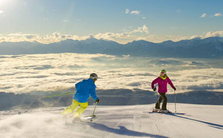 Two skiers carve down a freshly groomed slope above a sea of clouds, with sunlit Alpine peaks stretching across the horizon.