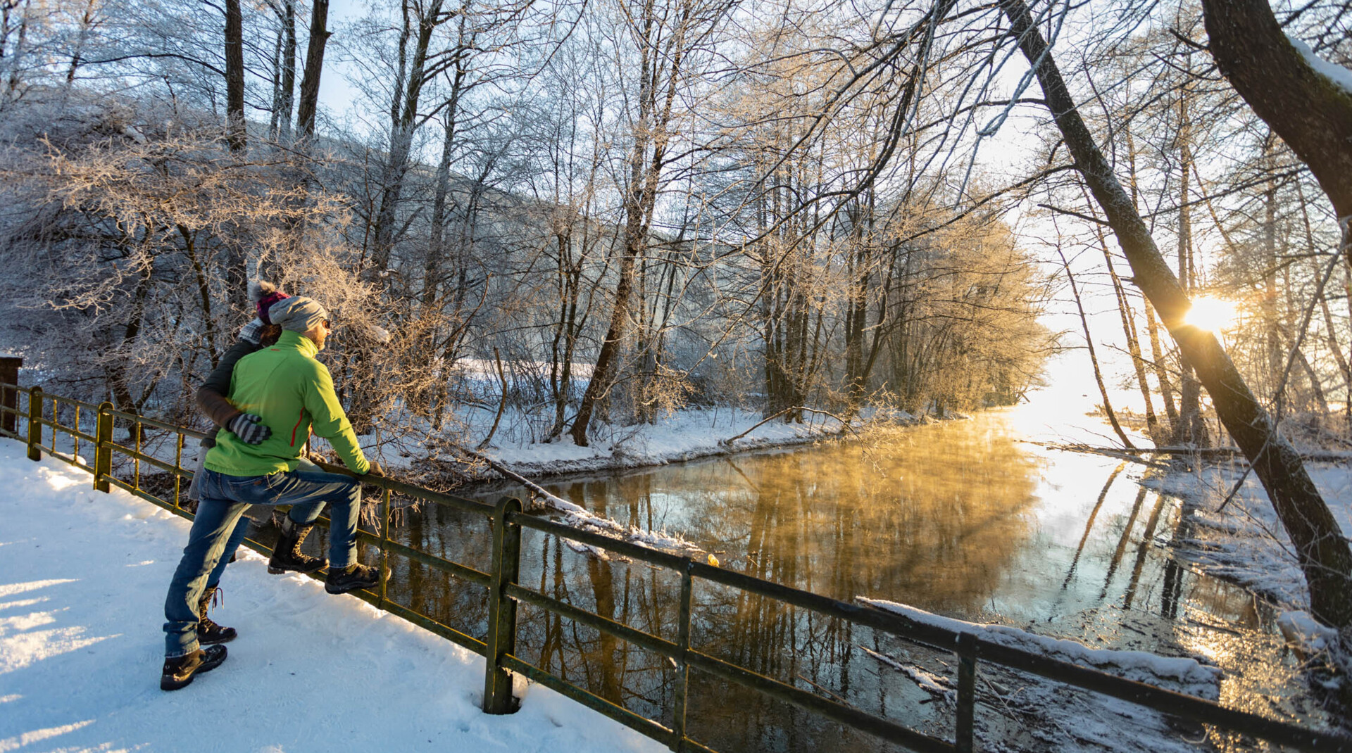 Two hikers on a snowy riverside walkway watch the sunrise lighting mist over a frosty stream.