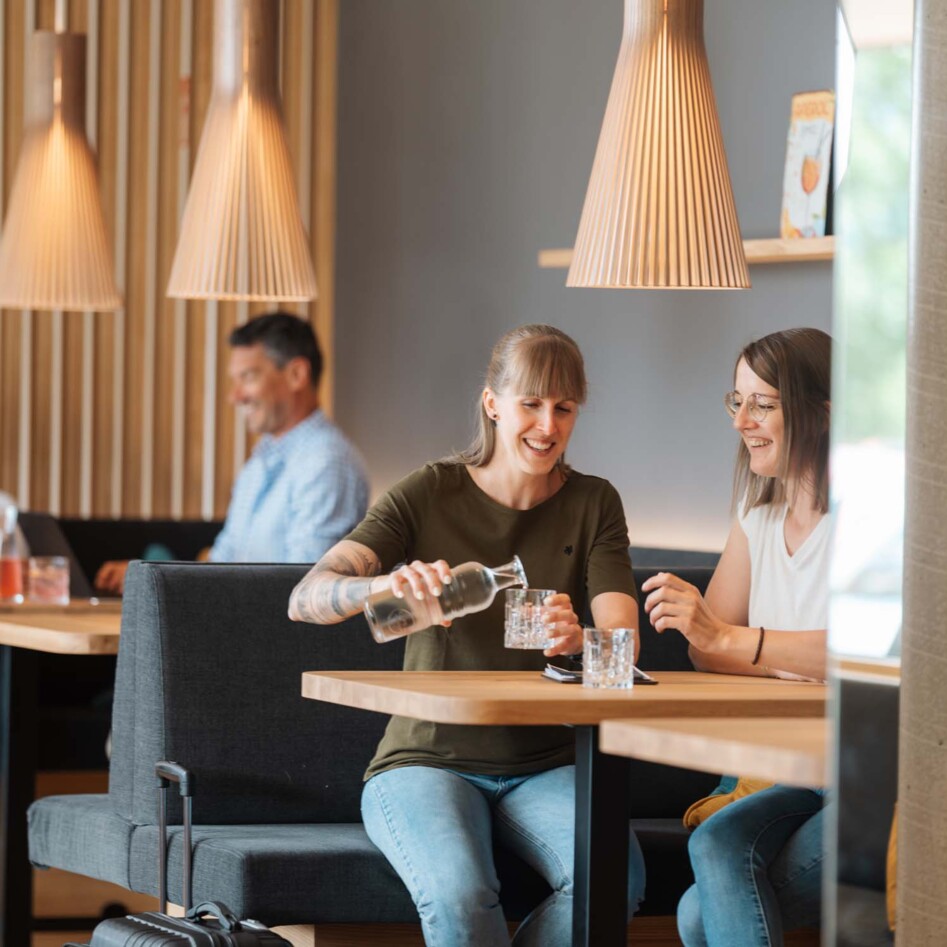 Zwei Frauen lachen, während sie Wasser einschenken. Im Hintergrund sitzen zwei weitere Personen an einem Tisch.