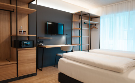 Modernes Hotelzimmer mit Doppelbett, Schreibtisch, Fernseher, und offener Kleiderablage. Helles, minimalistisches Design mit Holz- und Metallelementen.