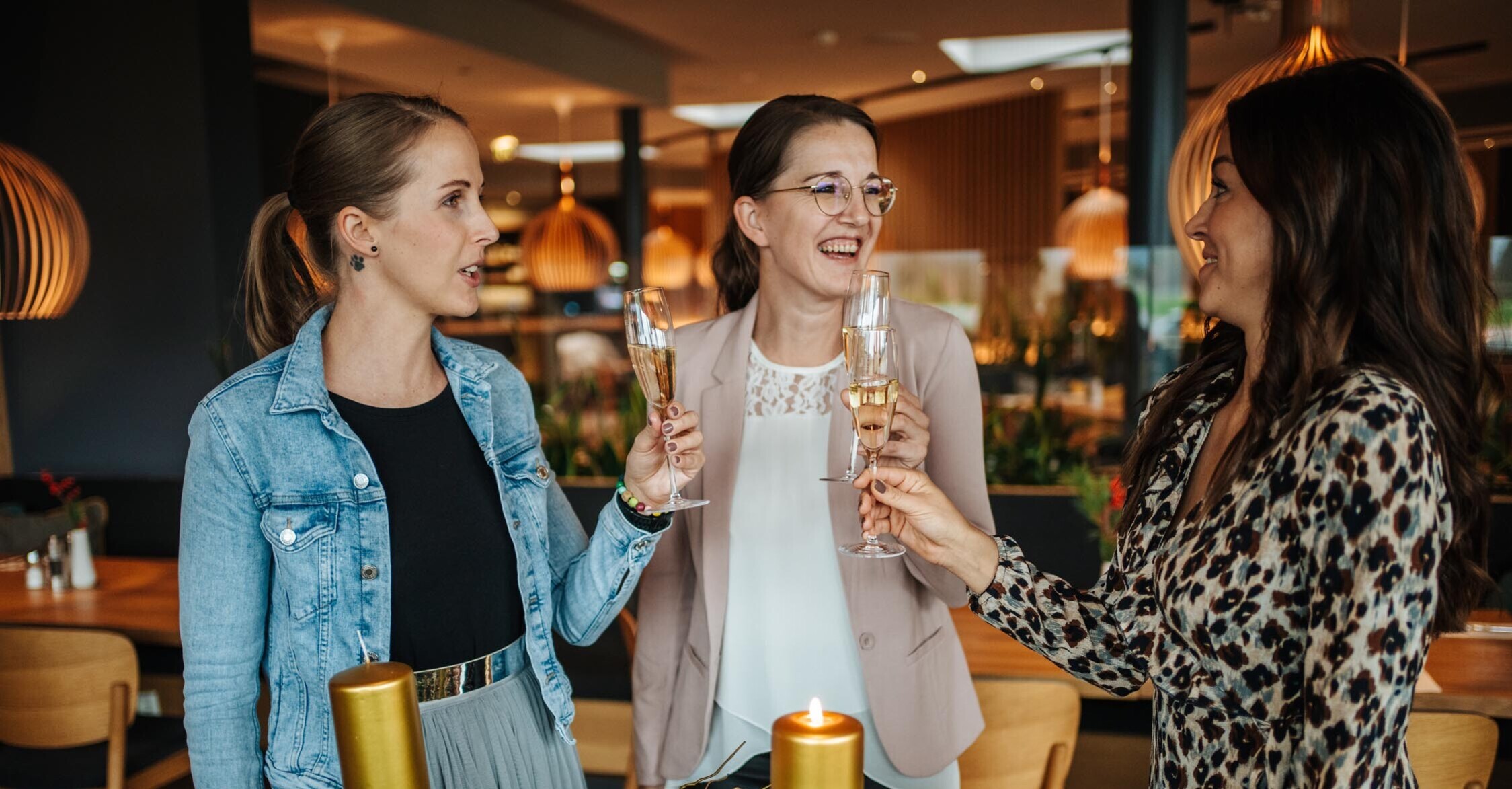 Drei Frauen stoßen mit Sektgläsern an, umgeben von festlicher Dekoration mit Kerzen und Tannenzweigen.