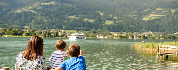 Eine Frau und zwei Kinder stehen an einem Seeufer und blicken auf ein Schiff, im Hintergrund ist eine Berglandschaft zu sehen.