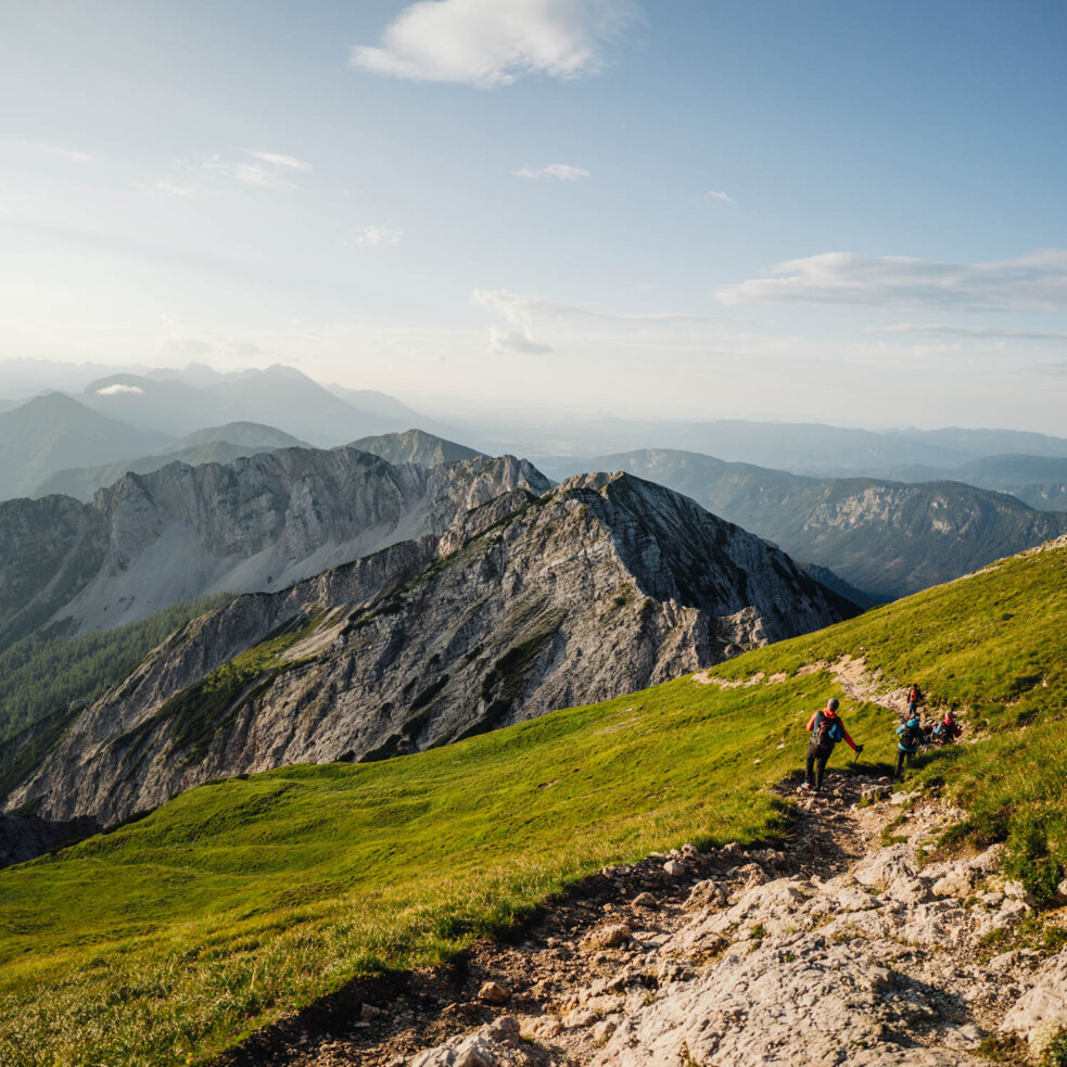 Wanderer auf einem Wanderweg am Mittagskogel mit Blick auf eine beeindruckende Berglandschaft bei sonnigem Wetter.
