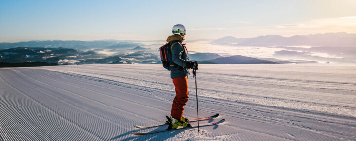 Ein Skifahrer steht auf einer frisch präparierten Piste mit Blick auf eine verschneite Berglandschaft bei Sonnenaufgang.