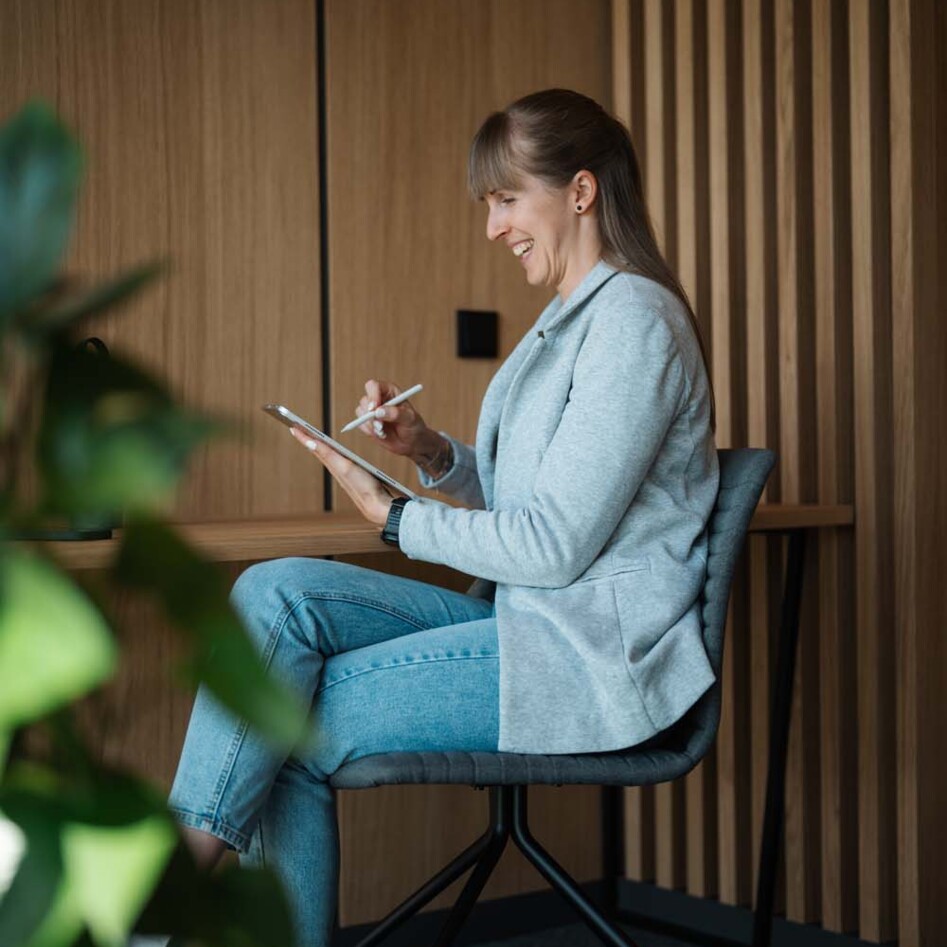 Eine Frau sitzt lächelnd auf einem Stuhl und schreibt auf einem Tablet. Im Hintergrund ist eine Holzwand zu sehen.
