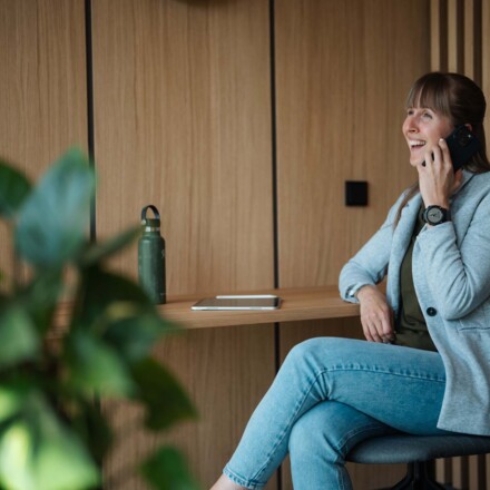 Eine Frau telefoniert lächelnd in einem Hotelzimmer, im Hintergrund sind Pflanzen und eine Trinkflasche zu sehen.