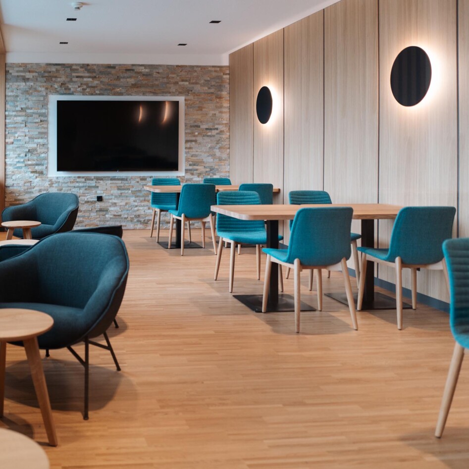 Restaurantbereich Hotel eduCARE mit blauen Stühlen, Holztischen und einem großen Bildschirm an der Wand.