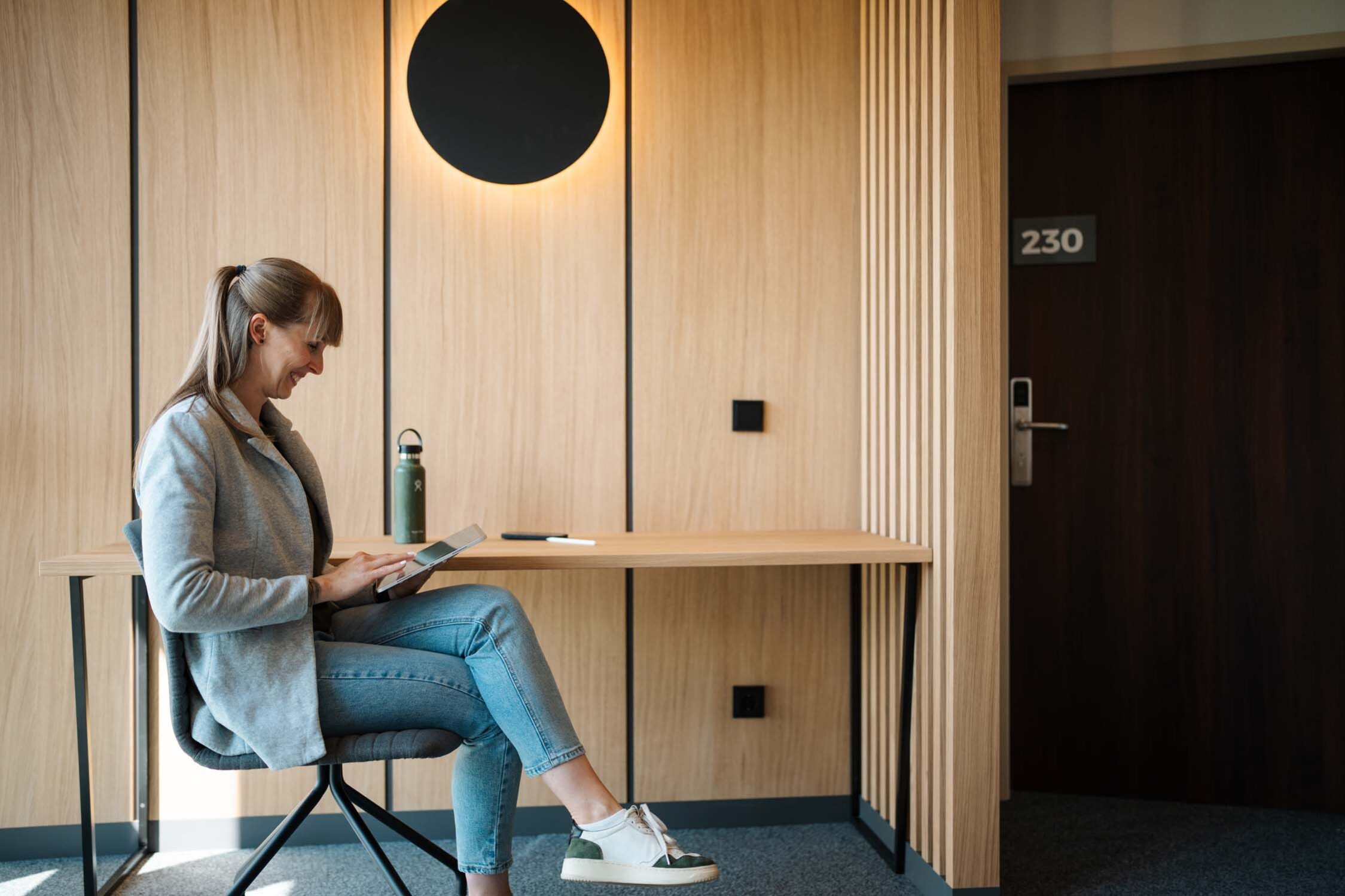 A woman sits at a wooden desk, smiling at a tablet. A water bottle and pen are placed on the desk.