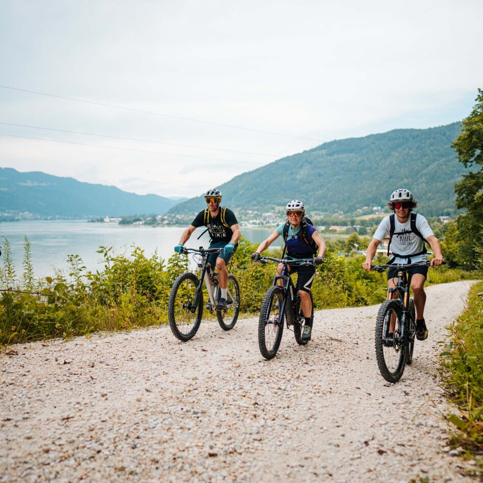 Drei Radfahrer fahren auf einem Schotterweg entlang eines Sees mit Bergblick.