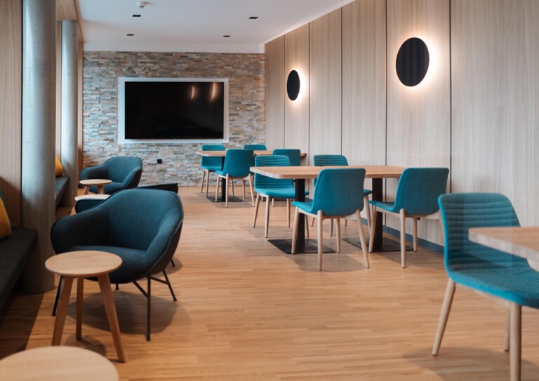 Restaurantbereich Hotel eduCARE mit blauen Stühlen, Holztischen und einem großen Bildschirm an der Wand.