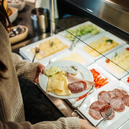 Eine Person bedient sich beim Frühstücksbuffet mit Wurst und Käse.