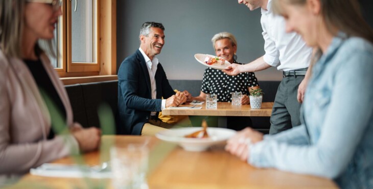 Personen sitzen im Restaurant, ein Kellner serviert die bestellten Gerichte.