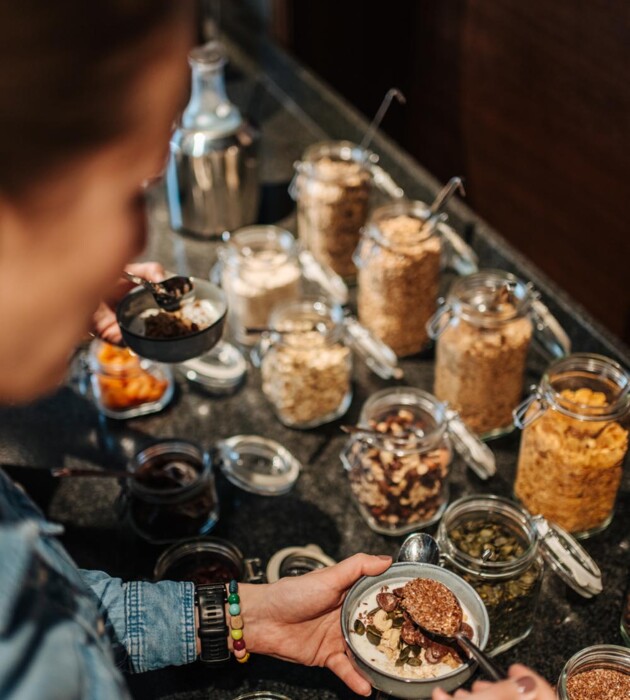 Eine Person bereitet eine Schüssel Müsli mit verschiedenen Zutaten aus Gläsern auf einem Tisch zu.