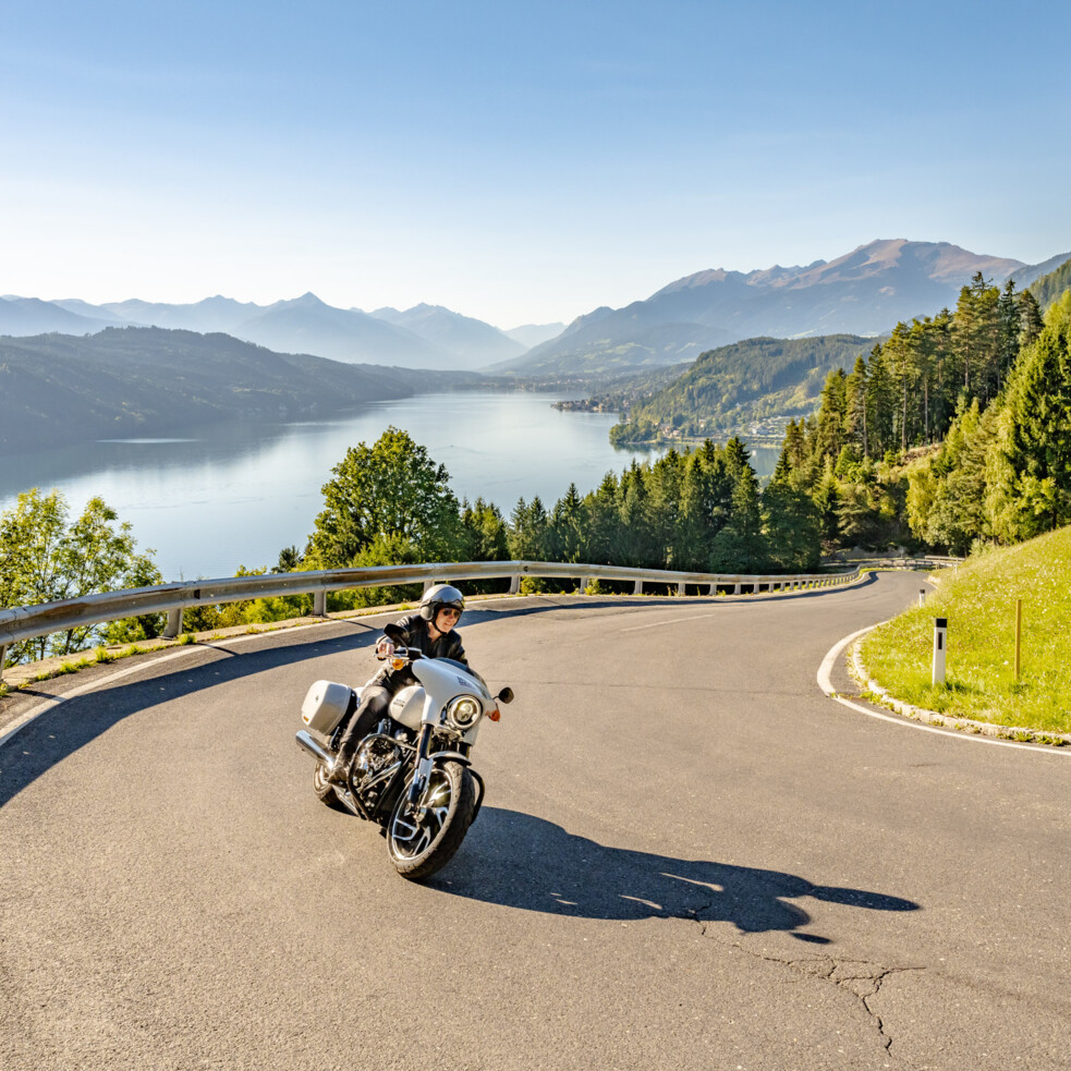Motorradfahrer auf kurviger Straße mit Blick auf See und Berge bei klarem Himmel.