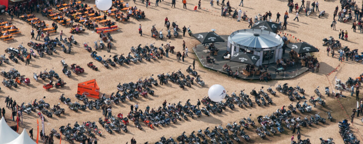 Luftaufnahme der European Bike Week am Faaker See mit vielen geparkten Motorrädern, Menschen und einem zentralen Pavillon, umgeben von Verkaufsständen und Sitzbereichen.