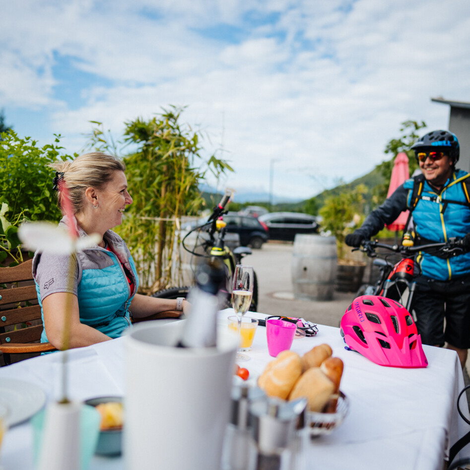Zwei Personen mit Fahrrädern unterhalten sich an einem Tisch in einem Gastgarten.