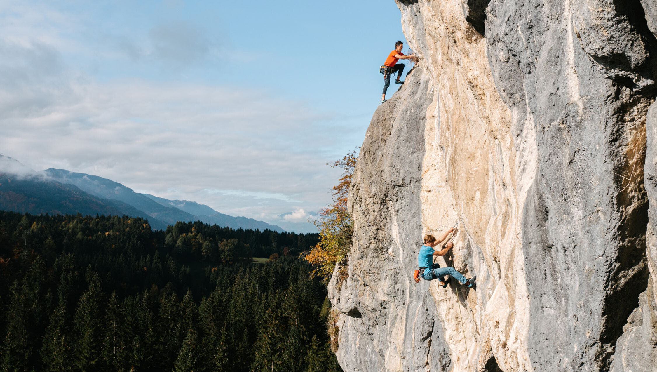 Zwei Kletterer erklimmen eine steile Felswand inmitten einer Wald- und Berglandschaft bei sonnigem Wetter.