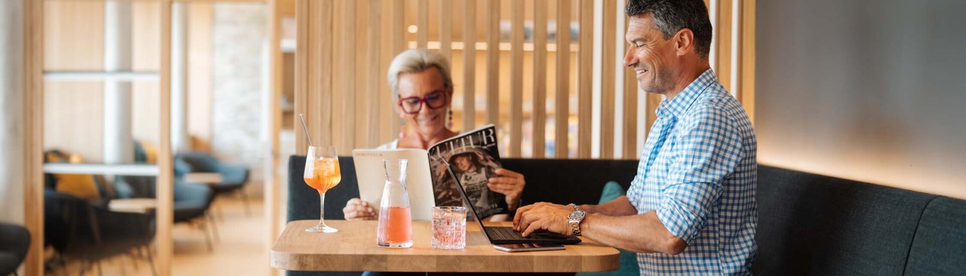 Ein Mann und eine Frau sitzen im Restaurantbereich. Die Frau liest eine Zeitung, der Mann arbeitet an einem Laptop.