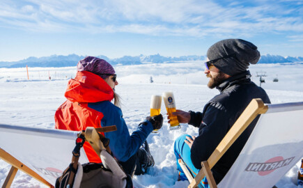 Two people in winter clothing clinking glasses on a snowy mountain, sitting on deck chairs with a scenic view of the Alps.