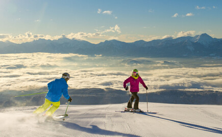 Zwei Personen beim Skifahren auf einer Piste mit Ausblicks auf eine Wolkendecke über dem Tal bei Sonnenschein