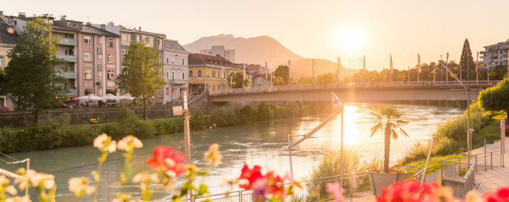 Sonnenuntergang über der Drau mit Brücke, umgeben von Blumen und Gebäuden, im Hintergrund Berge.
