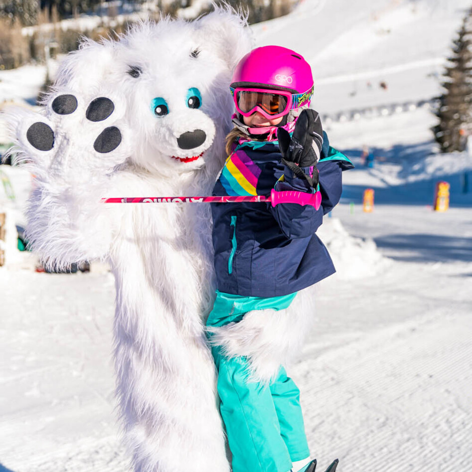 Child in ski equipment poses with the mascot Bino Bear on a snowy slope on the Gerlitzen Alpe.