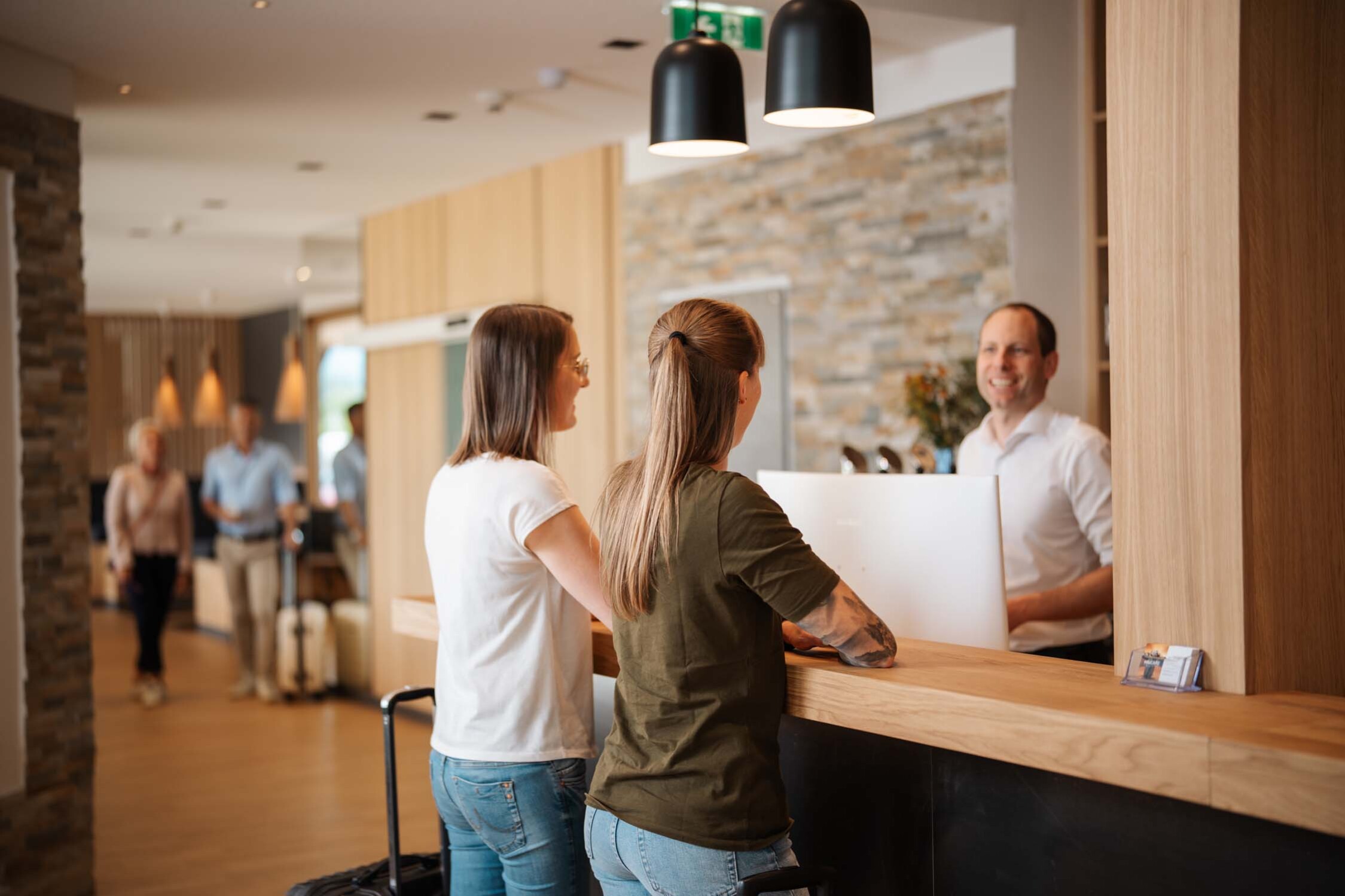 Two people at a hotel reception desk, engaging with a smiling receptionist. Others with luggage in the background. 