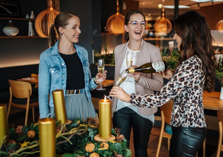Drei Frauen stoßen mit Sektgläsern im Restaurantbereich an und lachen dabei.