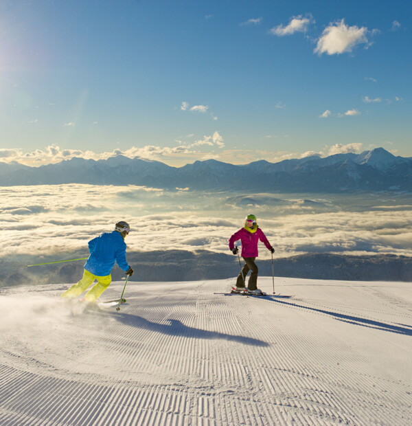Zwei Personen beim Skifahren auf einer Piste mit Ausblicks auf eine Wolkendecke über dem Tal bei Sonnenschein