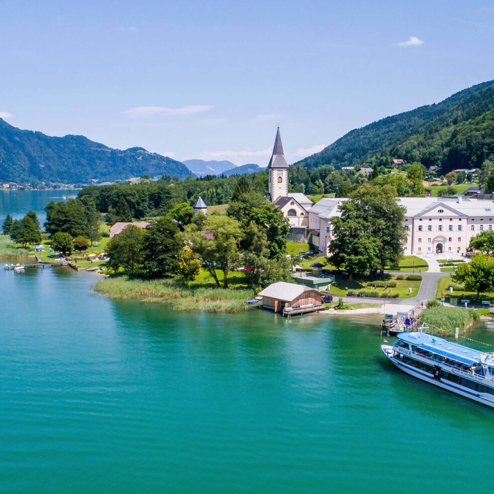 Luftaufnahme des Ossiacher Sees mit Boot. Am Ufer steht eine Kirche und weitere Gebäude.