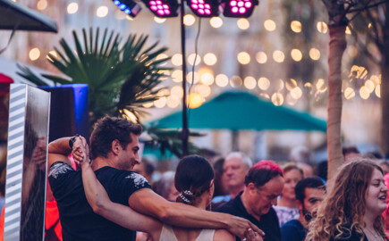 Menschen tanzen und genießen die festliche Atmosphäre bei Abendbeleuchtung auf einem belebten Platz.